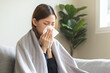 © KMPZZZ - Sick, coronavirus or covid-19, attractive asian young woman, girl have a fever, flu and hand in use tissues paper sneezing nose, runny while sitting on sofa, couch at home. Health care on virus person