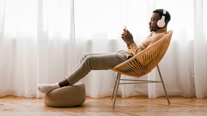 Aufkleber - Weekend. Relaxed Black Man In Wireless Headset Using Mobile Phone Listening To Audiobook Sitting On Modern Chair Against Window Indoor