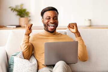 Canvas Print - Business Success. Excited Black Man Using Laptop Gesturing Yes Celebrating Great News Sitting On Couch Indoor. Copy Space