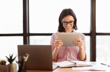 Poster - Surfing The Net. Happy young secretary in eyeglasses using digital tablet and smiling, sitting at desk in modern office