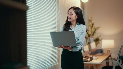 Poster - Businesswoman is standing in her home office, holding a laptop and looking out the window. She is wearing a blue shirt and black pants