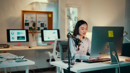 Poster - Asian businesswoman is sitting at her desk in a modern office, working late on her computer. She is recording a podcast using a professional microphone