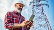 © angel_nt - A construction worker in a hard hat is focused on his smartphone while standing beside a tall transmission tower under a clear, blue sky
