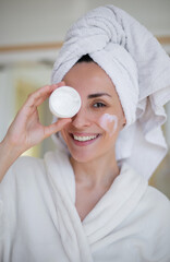 Wall Mural - Smiling woman with towel wrapped around her head holds a jar of cream over one eye and has a heart-shaped lotion mark on her cheek, symbolizing skincare, beauty, and self-care. Ideal for beauty ads