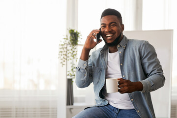 Poster - Coffee break in office. Cheerful black entrepreneur talking on phone and holding cup with hot drink in modern office space