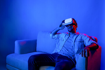 Poster - Happy man sitting at sofa while wearing VR goggle to watch funny movie. Caucasian person relaxed while looking at metaverse or virtual simulated world by using VR technology innovation. Deviation.