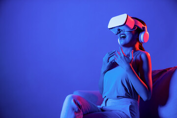 Smart female sitting on sofa surrounded by neon light wear VR headset connect metaverse, futuristic cyberspace community technology. Elegant woman excited and emotionally watch movie. Hallucination.