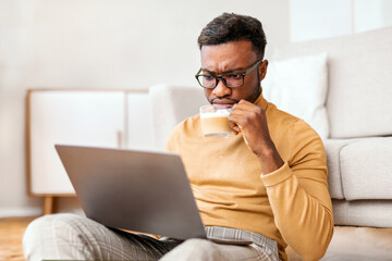 Aufkleber - Serious African American Guy Reading News On Laptop And Drinking Coffee Sitting On Floor At Home. Selective Focus