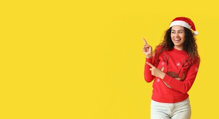 Canvas Print - Young African-American woman in Christmas sweater and Santa hat pointing at something on yellow background with space for text