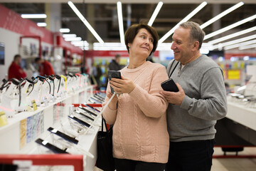 Mature couple, standing at the shelf with the goods, choose a new mobile phone in an electronics store