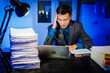 © Phushutter - A businessman works late into the night, facing a stack of papers on a black wooden desk. The blue lighting reflects the intensity of his overtime efforts in a busy office environment.