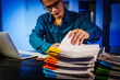 © Phushutter - Asian businessman works late into the night, focused on a stack of papers on his black wooden desk. The office is dimly lit with a blue glow, reflecting the busy atmosphere of overtime.