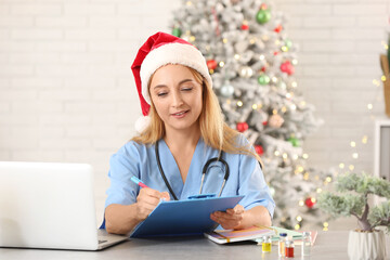 Canvas Print - Mature female doctor in Santa hat with clipboard at table in clinic