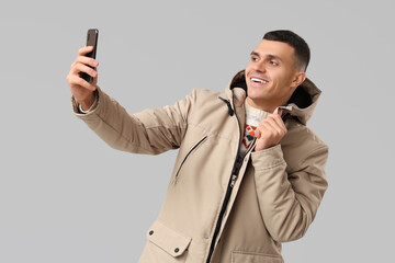 Wall Mural - Young man in stylish puffer jacket taking selfie on grey background