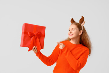 Young woman in reindeer horns with Christmas gift box on light background