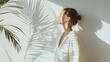© visual artstock - A woman stands with her eyes closed, bathed in the soft glow of sunlight streaming through a window, with the shadow of a palm tree cast on the wall behind her.