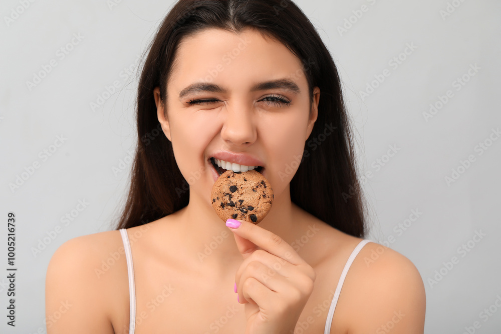 Beautiful young woman eating sweet cookie on grey background