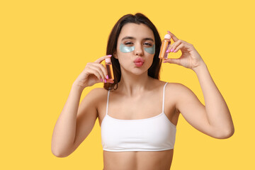 Sticker - Beautiful young woman with under-eye patches holding bottles of cosmetic products on yellow background