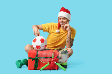 Sticker - Young happy man in Santa hat with badminton rackets, soccer ball and gift box on blue background