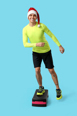 Poster - Young man in Santa hat training on aerobic stepper on blue background