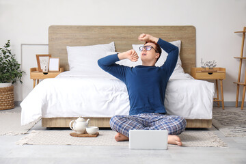 Sticker - Morning of handsome young man in stylish pajamas with teapot and laptop sitting at home