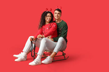 Sticker - Happy young couple in Christmas sweaters on sledge against red background