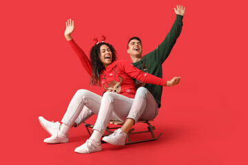 Sticker - Happy young couple in Christmas sweaters on sledge against red background