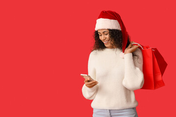 Poster - Beautiful young African-American woman in Santa hat with mobile phone and shopping bags on red background. Christmas celebration