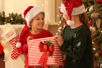 Sticker - Cute happy little children in Santa hats with gift box near Christmas tree on evening at home