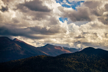 Breathtaking view of rugged mountains under a dramatic sky filled with clouds at sunset. Stunning landscape showcases towering mountains and lush forests, all bathed in warm light as stormy clouds