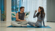 © Yuliia - Man woman sitting together on mats talking conversation drinking water holding glass break rest sport training athletes active lifestyle relaxing female male laugh positive dialog chatting wellbeing