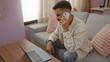 © Krakenimages.com - Hispanic man with beard working on laptop and talking on phone in living room apartment.