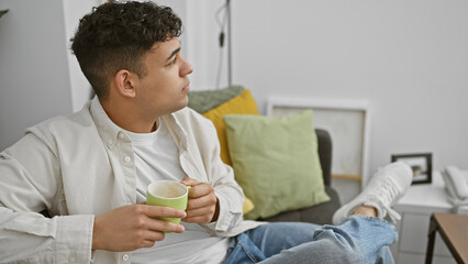 Wall Mural - A pensive young man with a cup sits relaxing in a modern living room, embodying casual home comfort.