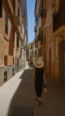 Woman walking through a narrow street in palma, mallorca, spain, wearing a hat and holding a wicker bag, under a clear blue sky in an outdoor setting surrounded by old buildings