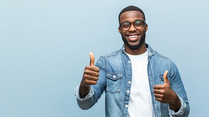 Cheerful young man in denim shirt and glasses giving enthusiastic thumbs up against light blue background, radiating positivity and confidence.