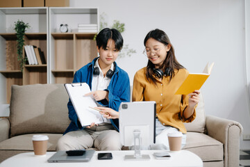 Two young Asian student studying at the college library, sitting at the desk, using a laptop computer, tablet and headphones having a video chat.