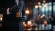 © Elmira - A butler stands tall in a polished black suit, holding a brass bell with one gloved hand, ready to summon service in an upscale environment filled with soft lighting