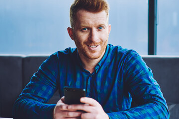 Portrait of cheerful hipster guy with modern mobile phone in hands looking at camera during break for studying in university campus, positive smiling male student preparing to college examination