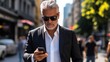 © Denis - an elegant man wearing a suit, sunglasses, with a serious expression, gray hair, looking at his cell phone as he walks through the streets of the busy city.