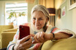 © Marko Geber - Smiling senior woman using smartphone on couch at home