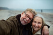 © Marko Geber - Smiling senior couple taking a selfie on beach during winter