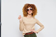 © SHOTPRIME STUDIO - Stylish woman with curly hair wearing sunglasses and a beige sweater against a light background, smiling confidently and posing with one hand on her hip