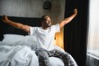 © Serhii - Good Morning. Happy Black Man Waking Up And Stretching Arms With Smile, Relaxed After Great Rest, Sitting In Bed In Modern Bedroom Interior. Start Of New Day, Rest And Wellness Concept