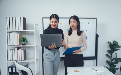 Businesswomen Collaborate on Project: Two smiling businesswomen stand in a modern office, engrossed in a collaborative project, with a laptop and folder, showcasing teamwork, communication.