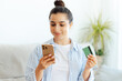 © Kateryna - Positive attractive brunette woman, sits on a sofa in a living room, holds smartphone and credit card, enter card information for online payment in a mobile app, making online shopping, smiling