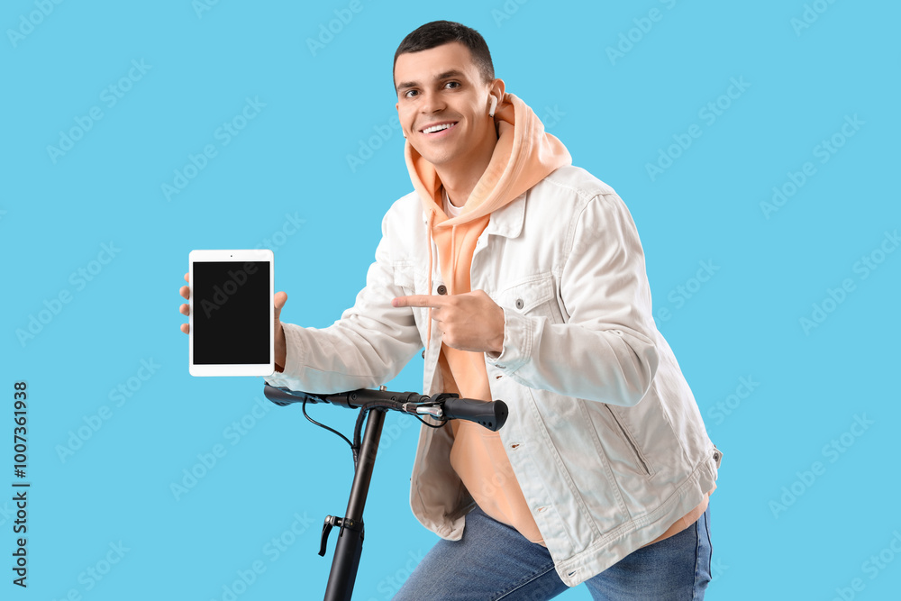 Young man with electric scooter pointing at tablet computer on blue background