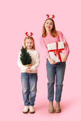 Wall Mural - Cute little girl in reindeer horns with small Christmas tree and her mother holding gift box on pink background