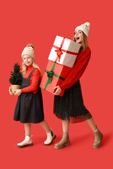 Wall Mural - Cute little girl with small Christmas tree and her mother holding gift boxes on red background