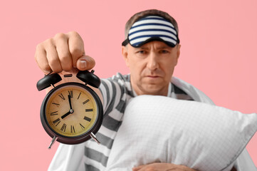 Poster - Handsome mature displeased man in stylish pajamas with pillow and alarm clock on pink background