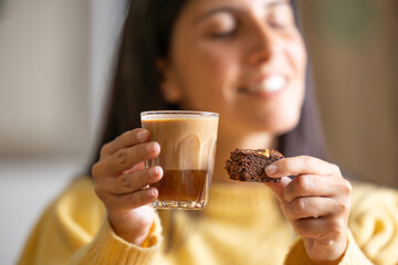 Happy young woman smiling enjoys her breakfast or lunch recovering energy with a delicious caramel coffee and eats a chocolate cupcake, close-up in a specialized pastry cafe on wooden table